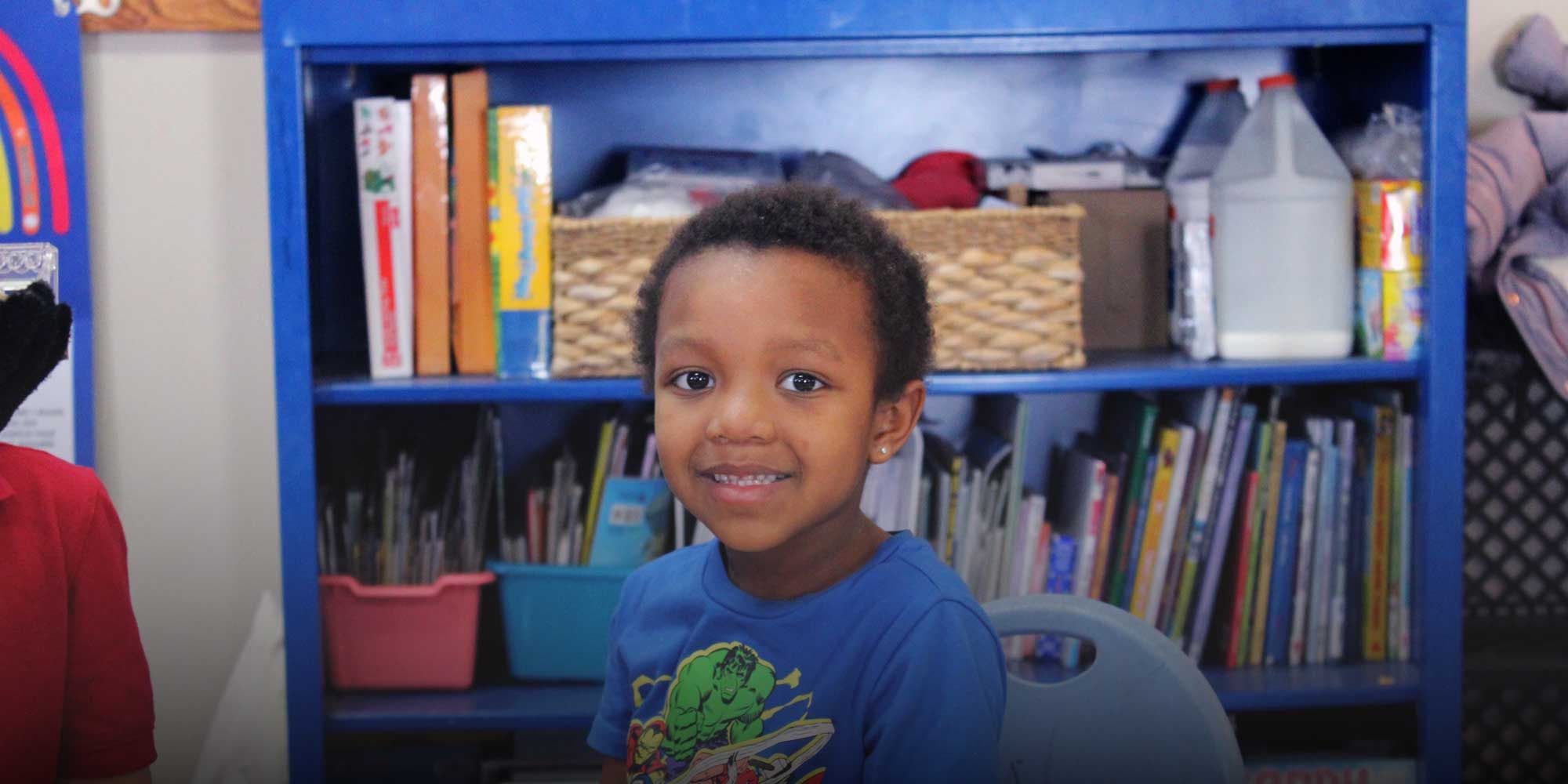 Smiling student in a blue shirt in front of a classroom bookshelf.