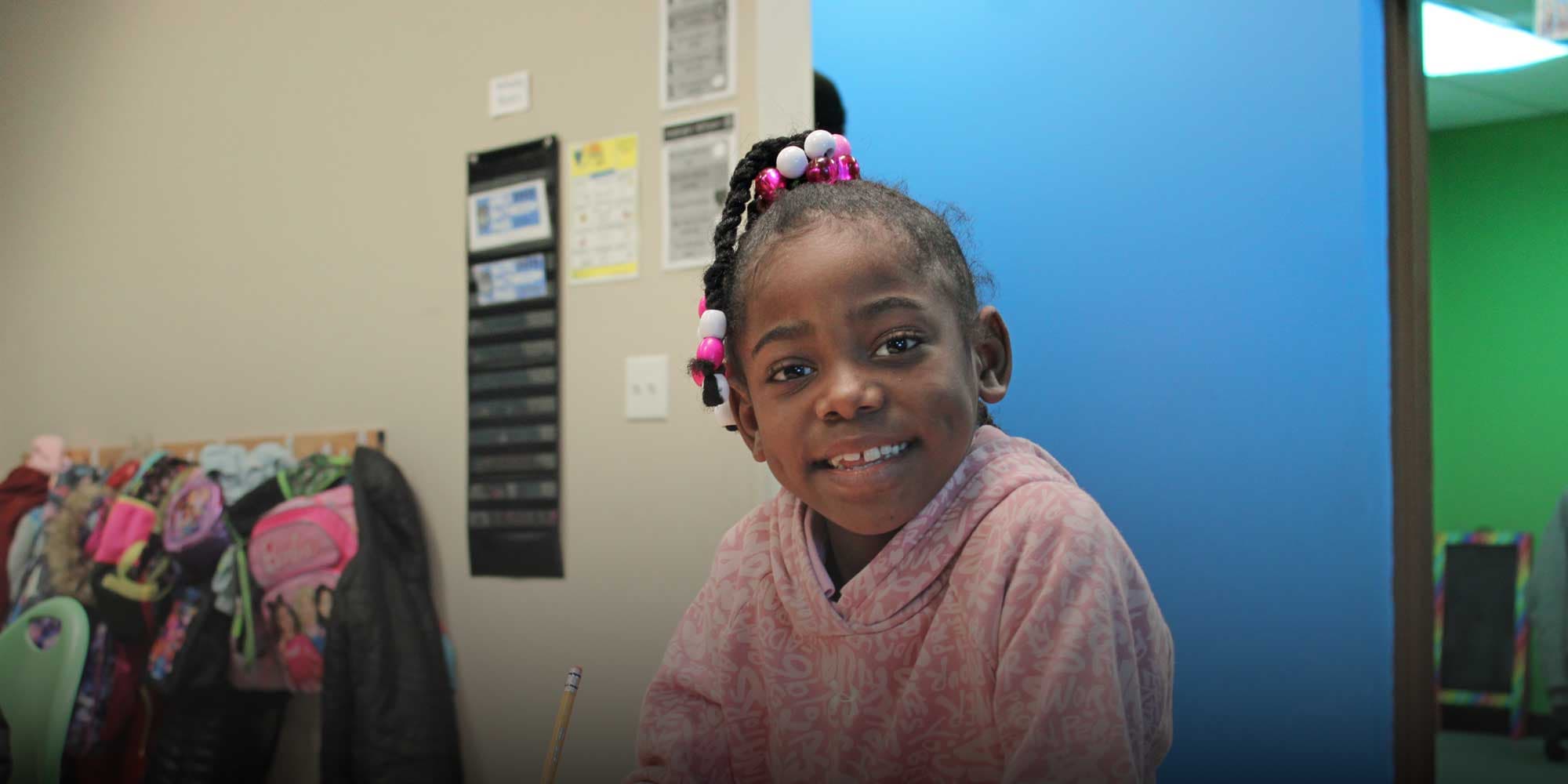 Smiling student with beaded braids and a pink sweater in a classroom.